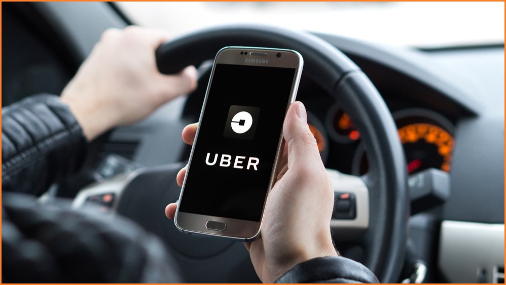 Man driving holding steering wheel and mobile phone with Uber logo on screen