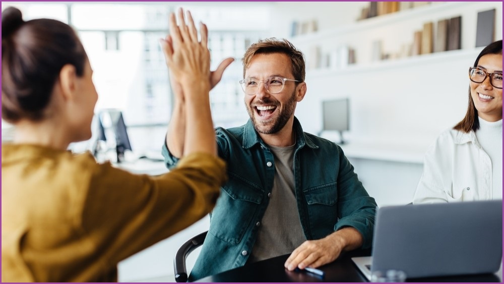 Two office workers high-five each other while a third worker watches on.