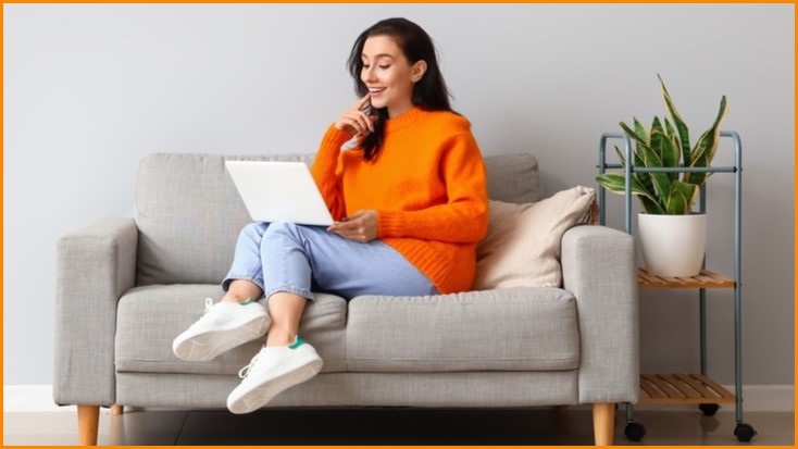 Woman in orange shirt working on a laptop while sitting on a couch.