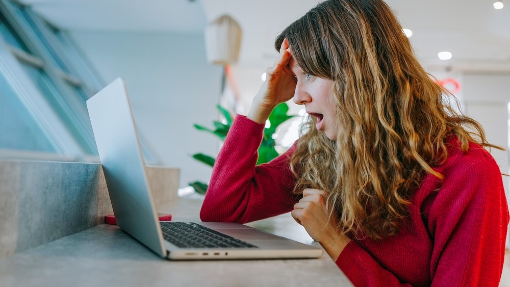 Woman in red jumper looking in shock at her laptop screen.