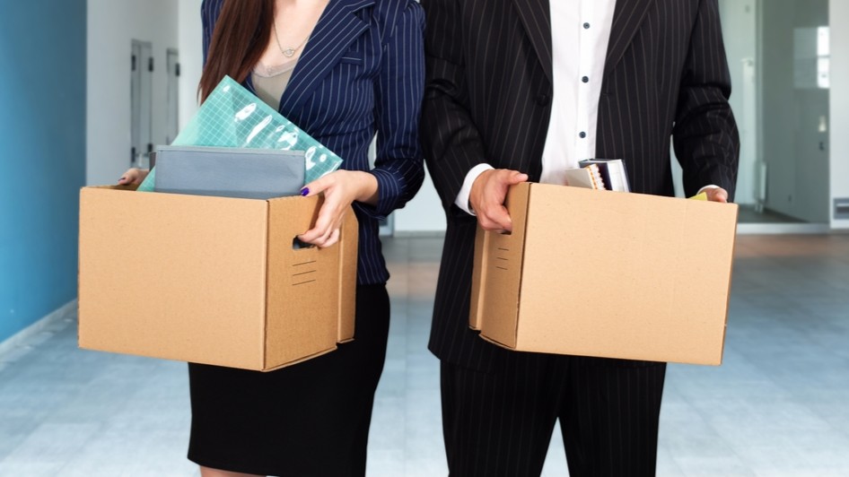 Two people leaving a workplace, each holding a cardboard box containing their possessions.