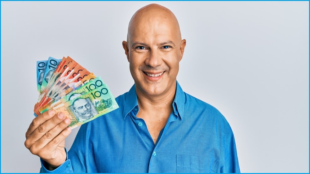 A smiling bald man in a blue shirt holding a handful of Australian dollars.