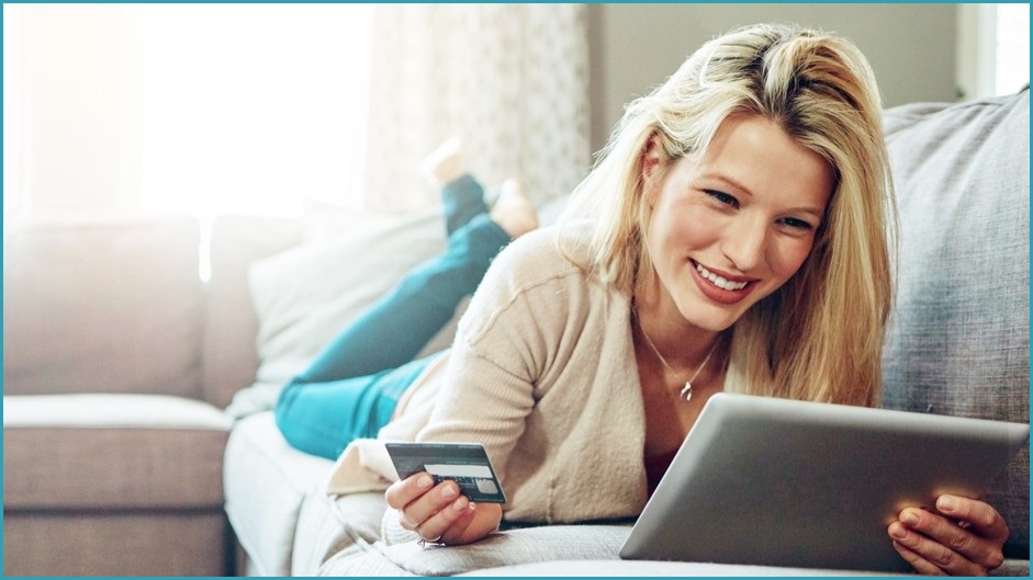 Smiling blonde woman propped up while laying down on a couch, holding a credit card in one hand and looking at a tablet screen.