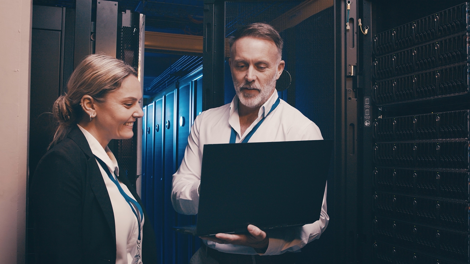 A medium shot of a man holding a laptop and a woman looking at it, standing in the hallway of a server room.