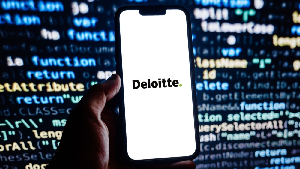 Person holding a phone with the word 'Deloitte' on the screen aganist a backdrop of random text on a computer screen.