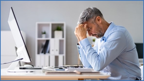 Old man sitting at a computer, looking down, resting his forehead on his hands.