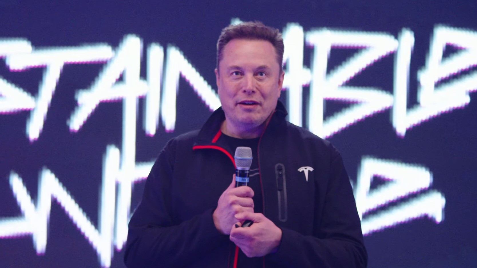 A medium shot of Elon Musk holding a microphone and speaking.