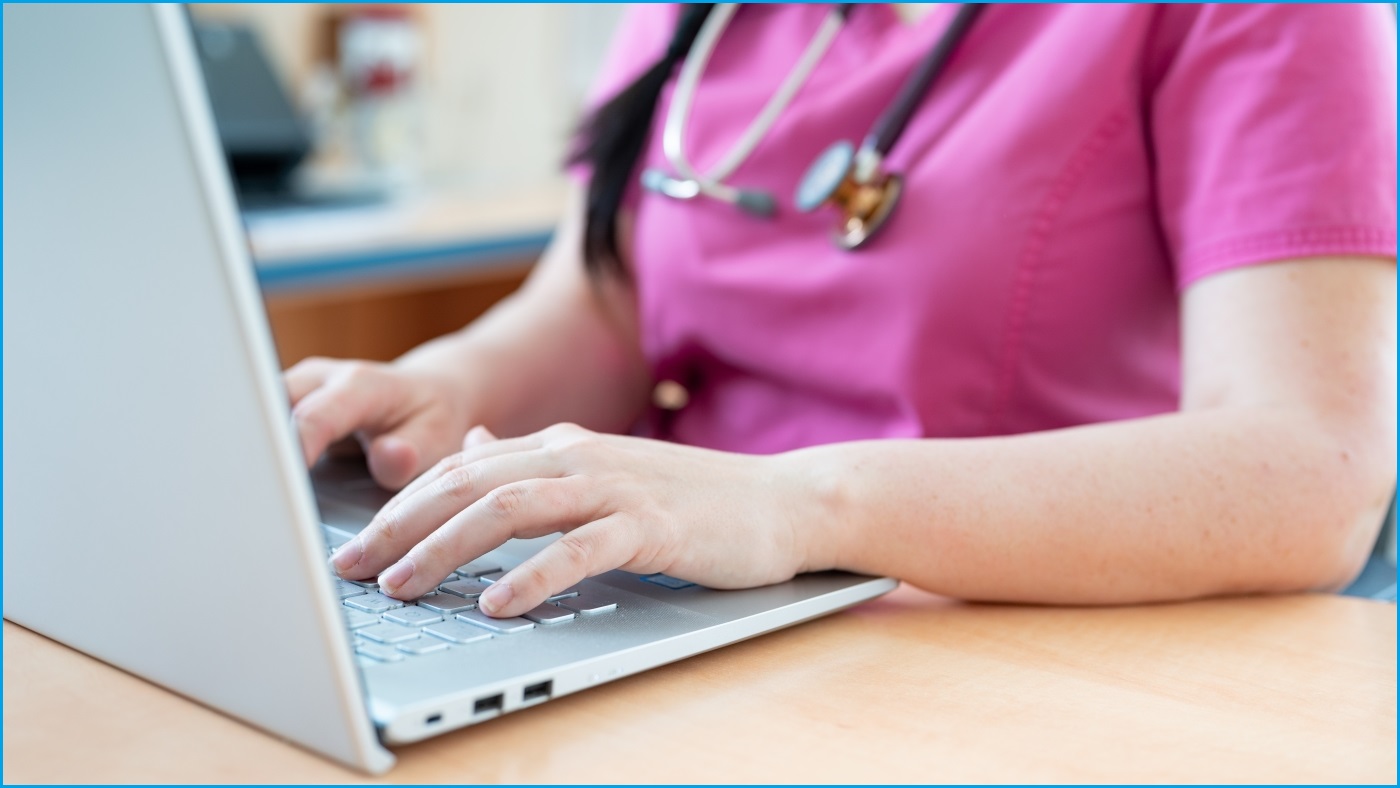A close up of a female doctor using a laptop, and wearing a stethoscope around her neck.