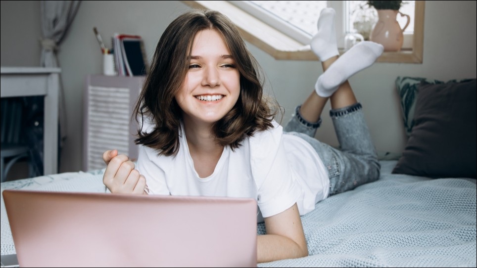 Teenage girl lying face down on a bed working on her laptop.