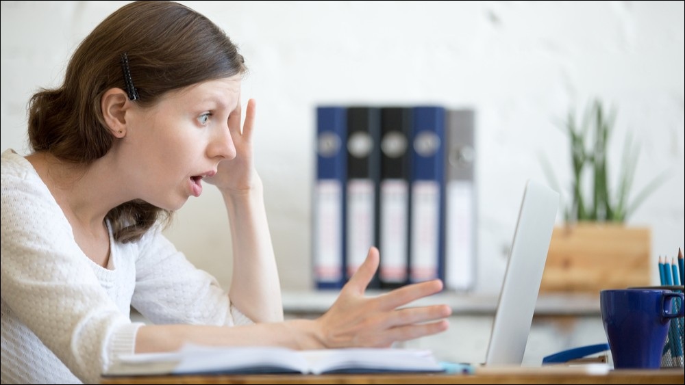 A medium shot of a woman looking at her laptop in shock, with mouth wide open.