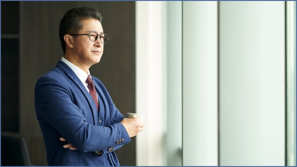 Older male executive looking out a window, in a contemplative stance.