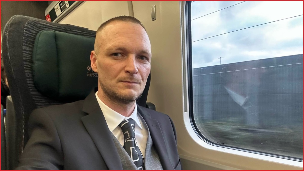 A medium shot of a man with short hair and wearing a suit sitting on a moving train and looking at the camera.