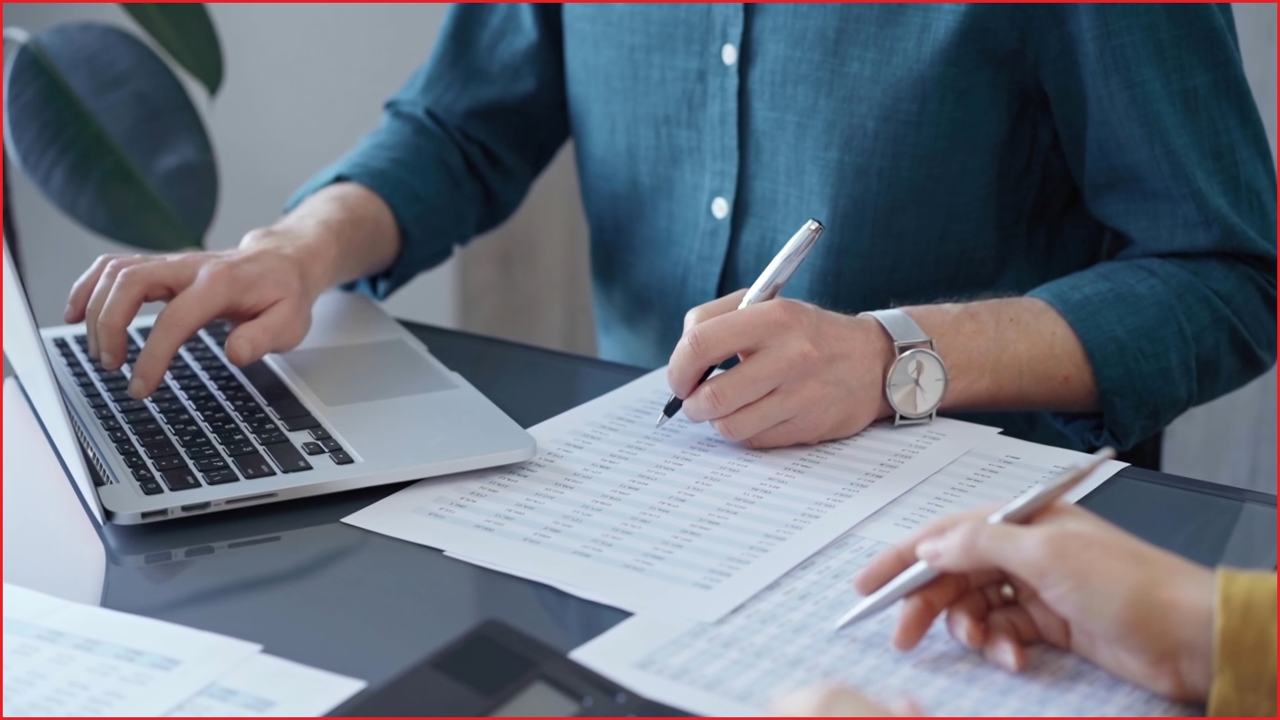 A close up of a person's hands, one using a laptop, and the other writing with pen and paper.