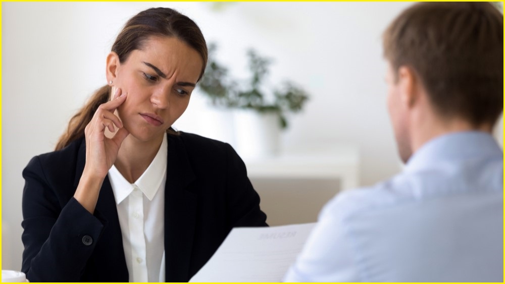 A medium shot of a woman in business attire looking wth confusion at a piece of paper, as a man sits opposite her at a table.