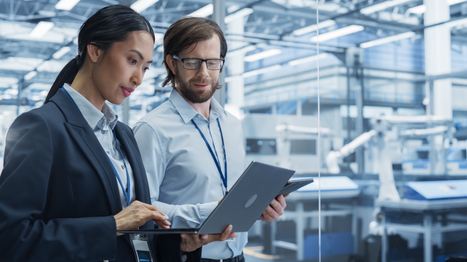 A medium shot of a man and a woman wearing business attire and holding a laptop and a tablet, while walking through a modern factory.