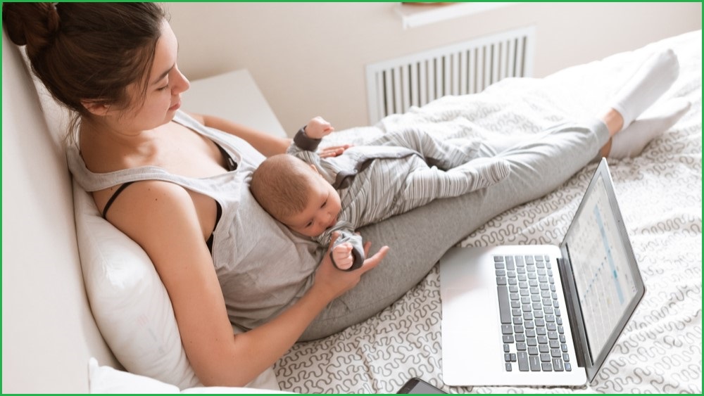 Woman sitting up in bed with baby laying on her lap and laptop nearby.