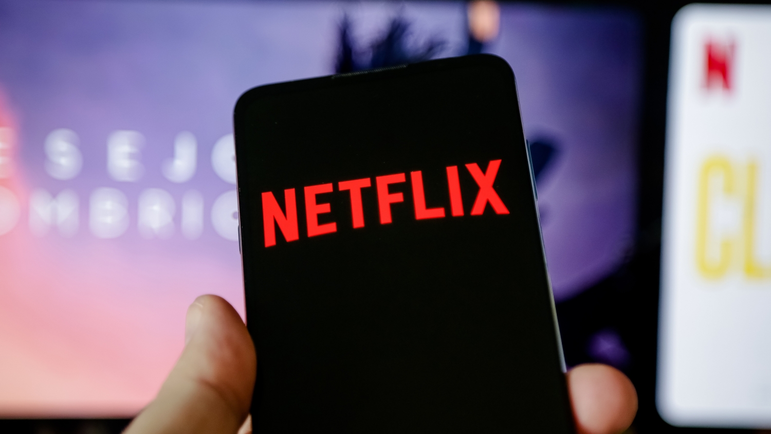 A close up of a smartphone showing the Netflix logo. In the background, a TV displaying a Netflix menu.