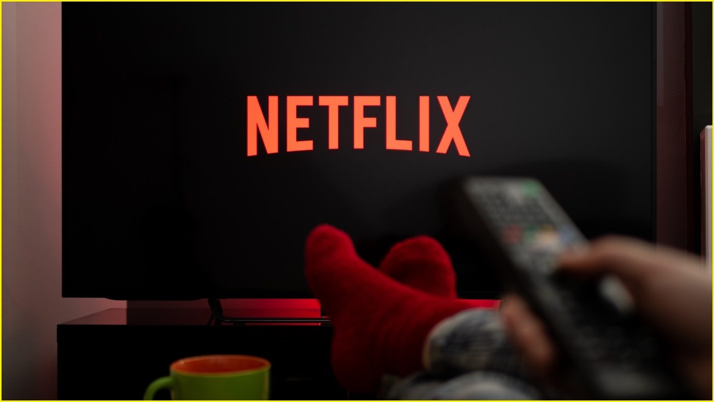A close up of a hand holding a TV remote, with a TV in the background displaying the Netflix logo.