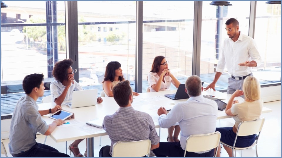 People sitting around a work table with one person standing up and speaking.