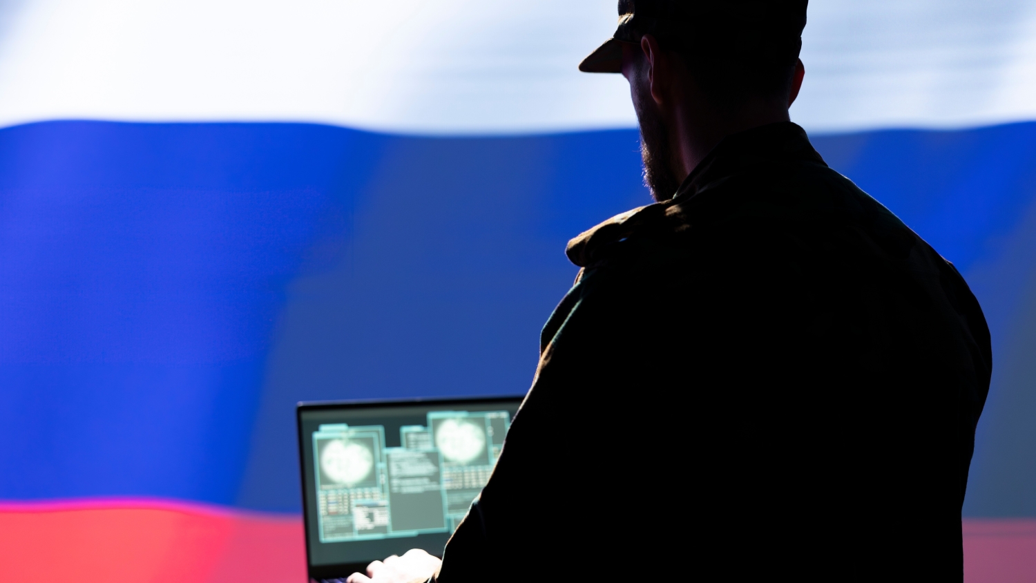 A medium shot of the back of a male figure standing and using a laptop. in the bacground is a large Russian flag.