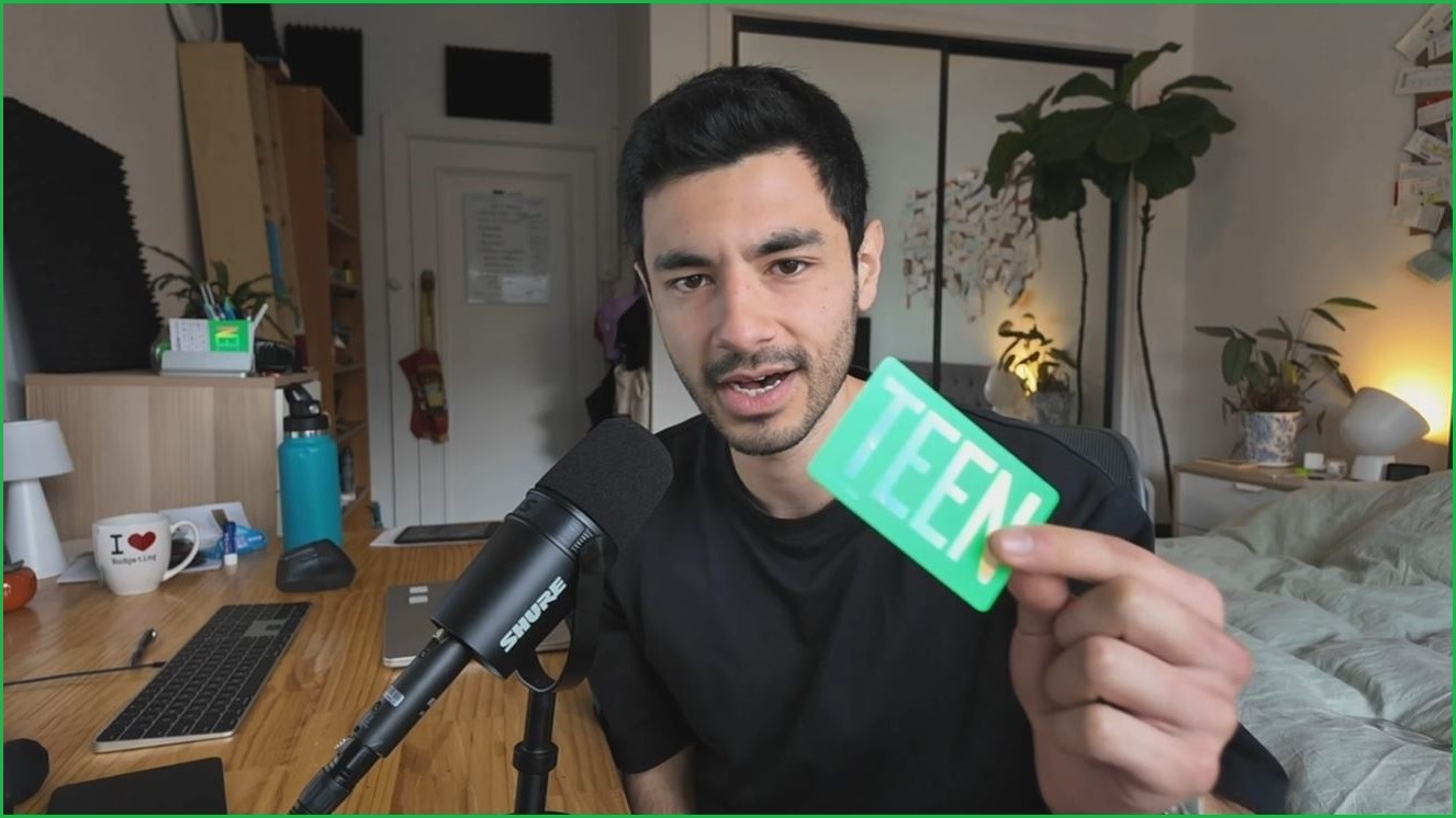 A medium shot of a man sitting at a desk in a bedroom speaking into a microphone and holding up a gift card with the word 'Teen' on it.