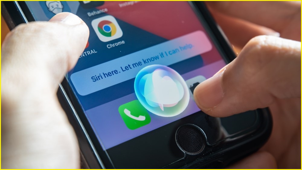 A close up of a person's hands holding and using an iPhone, which is displaying the Siri bubble on screen.