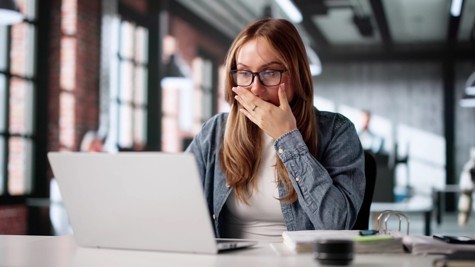 Woman gasping, holding hand over her mouth, looking at a laptop screen.