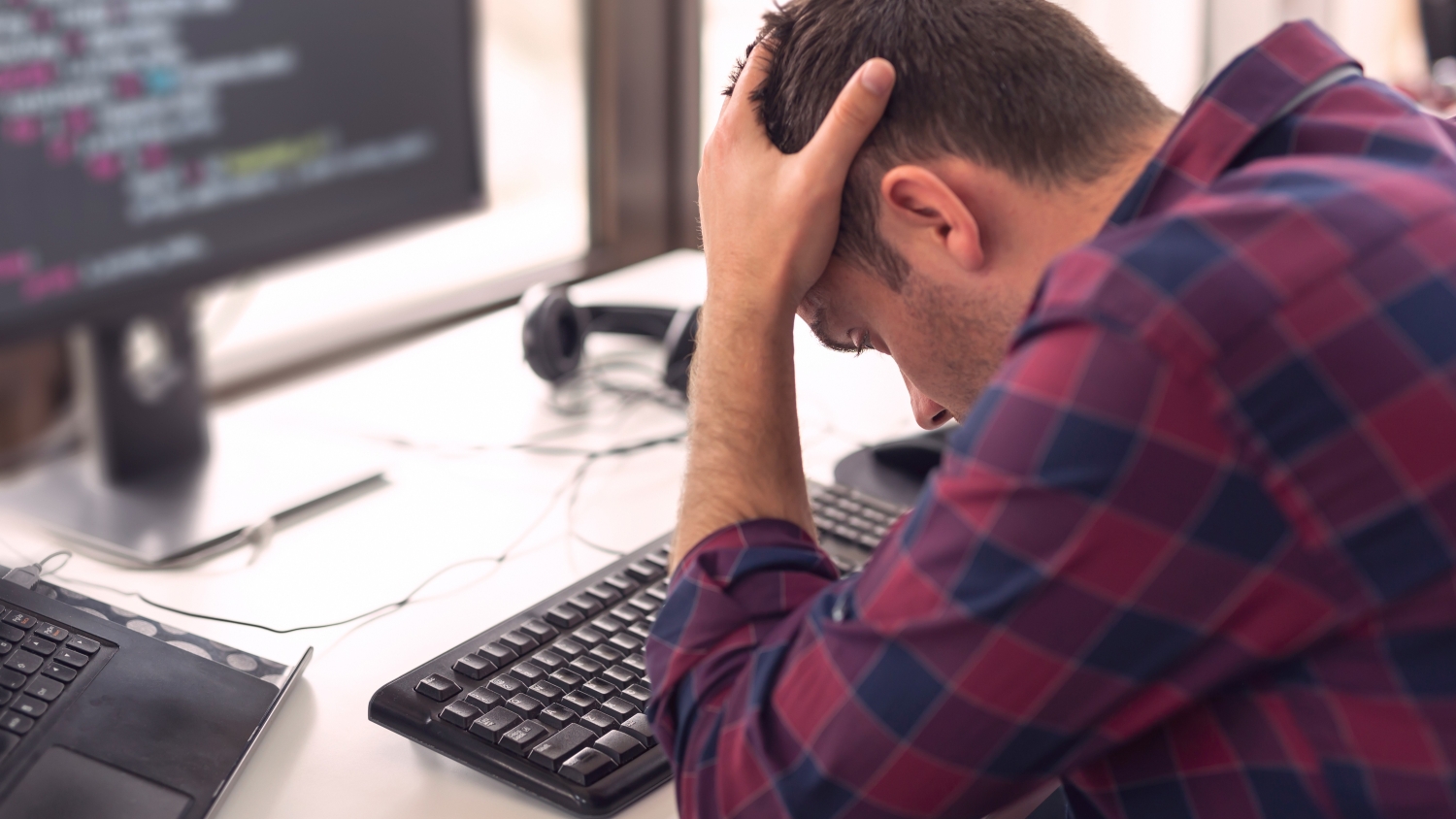 A close up of a man at a desk with his head in his hands, leaning on the desk, in front of a computer