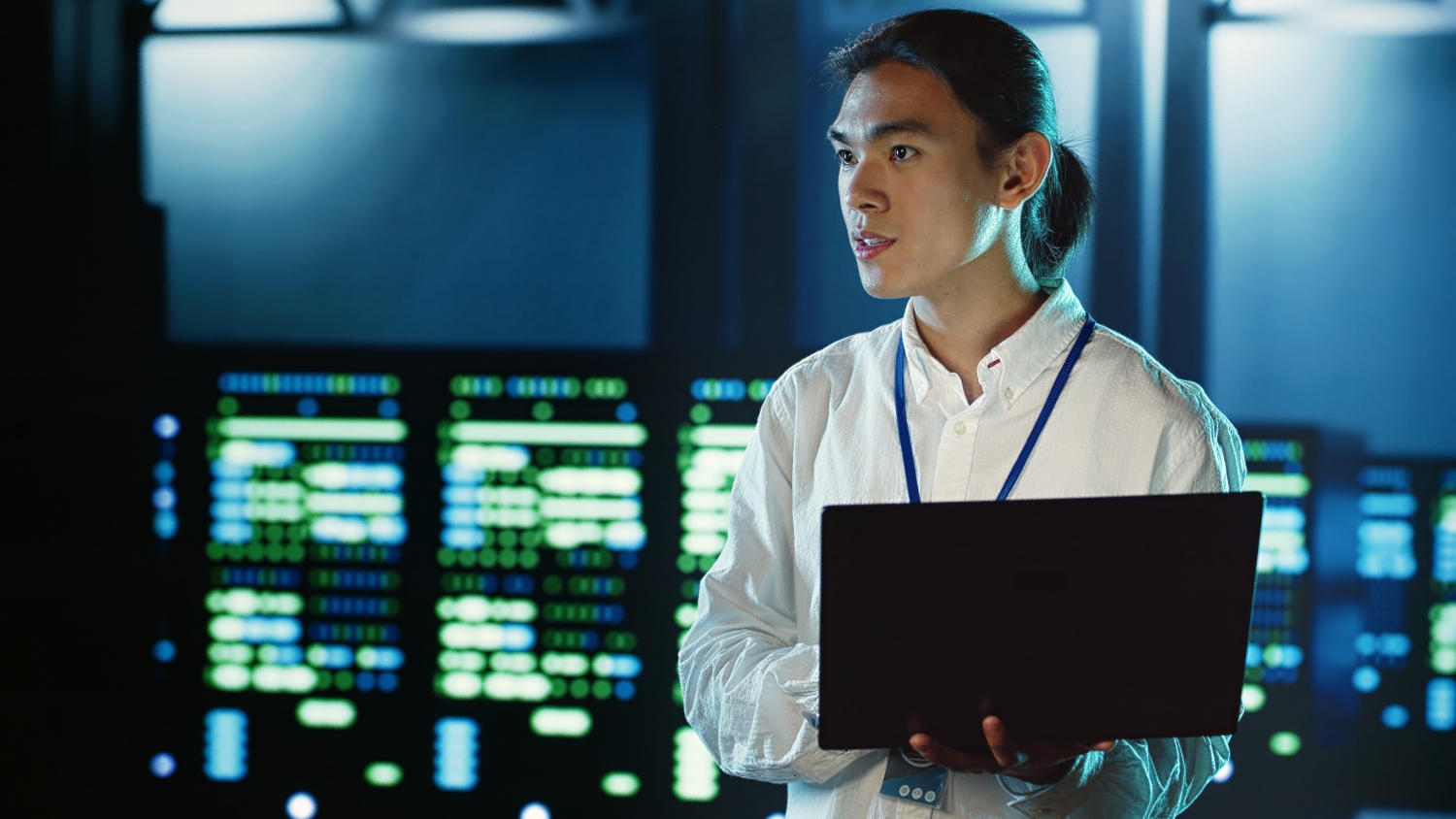 A medium shot of a man holding a laptop and standing in a server room.