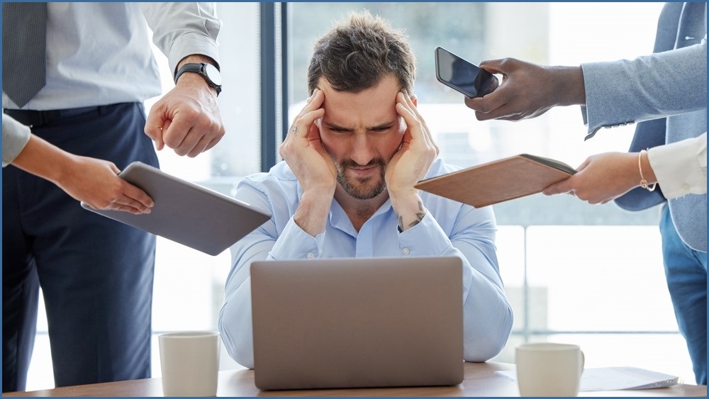 Man looking at a laptop screen holding his temples, looking stressed, as people thrust different technology at him.