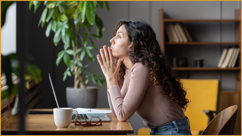 Thoughtful woman with elbows on the desk, palms together, staring upwards into the distance.