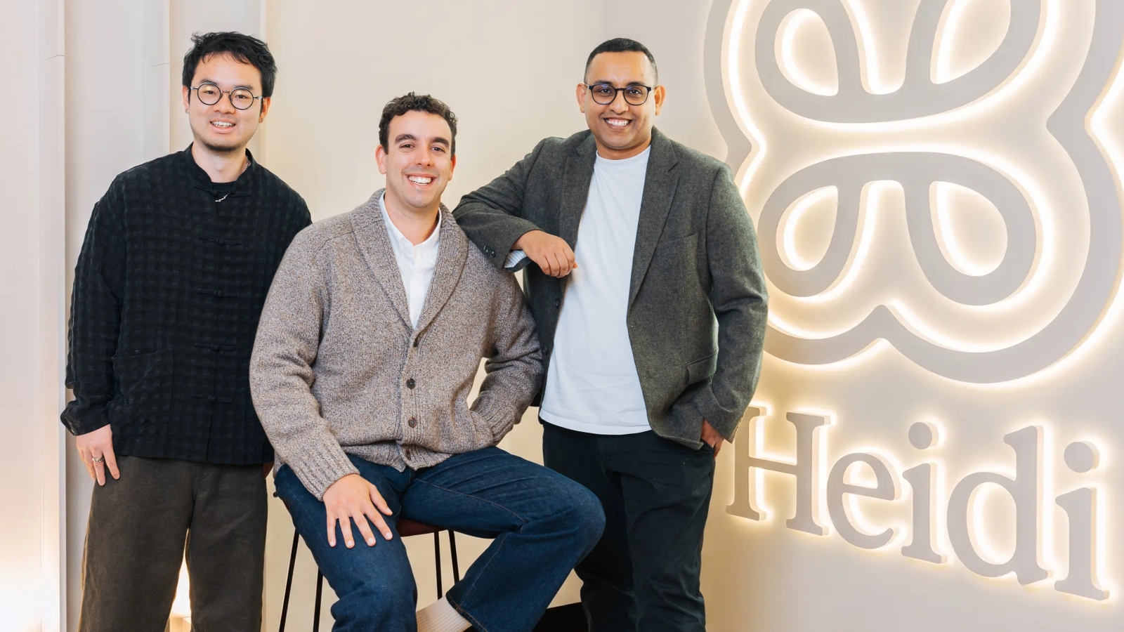 Three men smiling at camera, next to a wall with the Heidi AI logo on it.