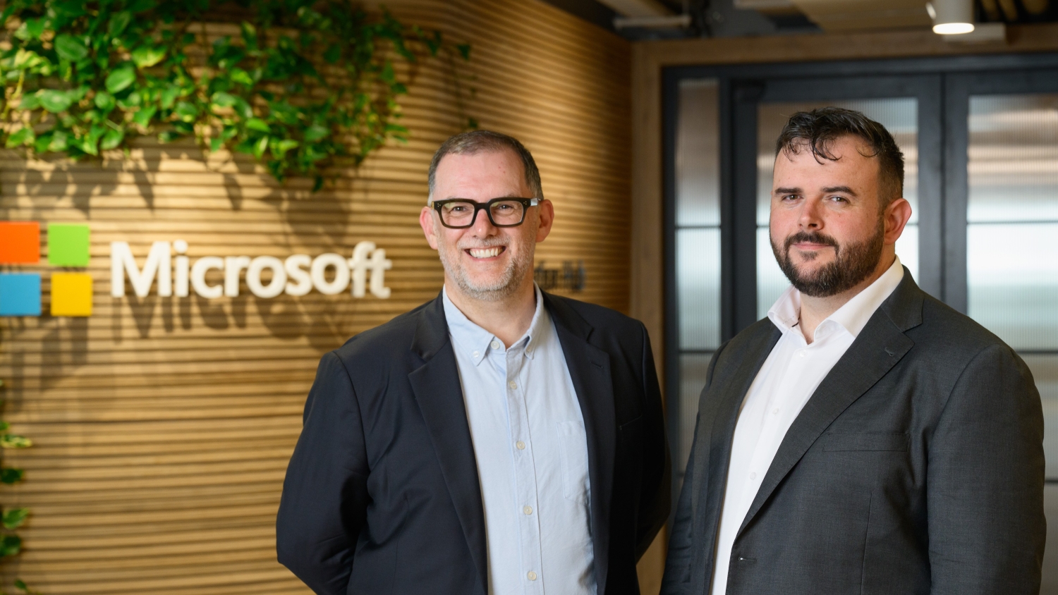 Two men in suit jackets stand and smile near a wall which bears the Microsoft logo.