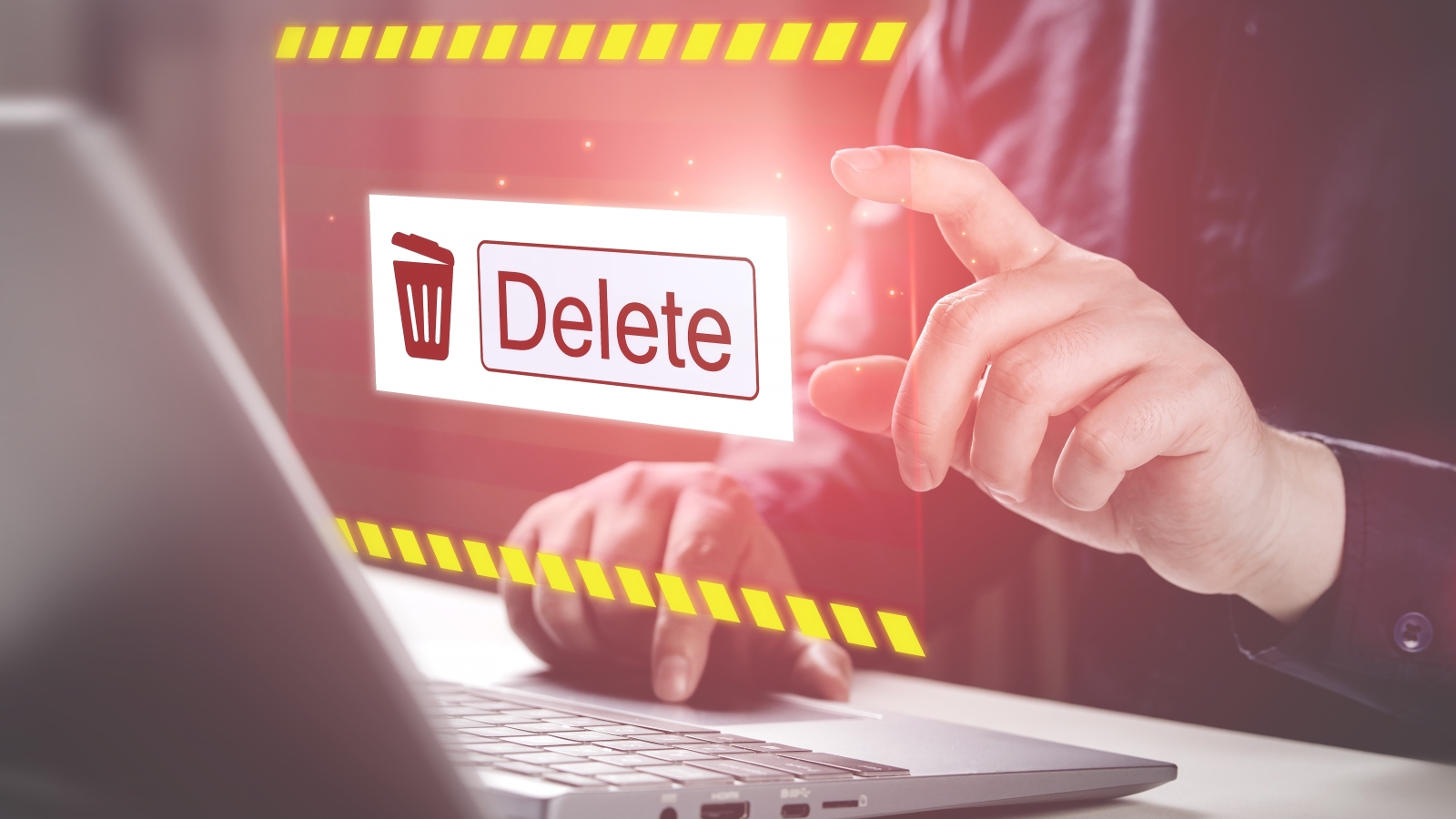 A closeup of a person's hands using a laptop. Above the laptop flots a large computer icon of a trash bin and the word 'Delete'