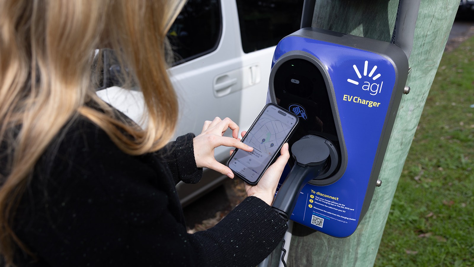 A close up of a woman using a smartphone while standing at a kerbside AGL EV charger.