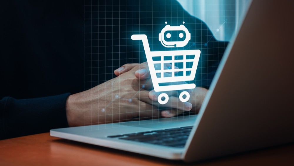 Person sitting with folded hands at a laptop with an image of a shopping trolley with a robot head floating mid-air.