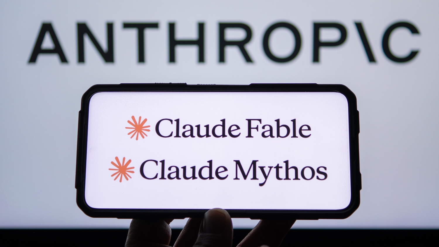 A closeup of a smartphone showing the Claude Fable and Claude Mythos logos. In the background, the Anthropic logo on a wall