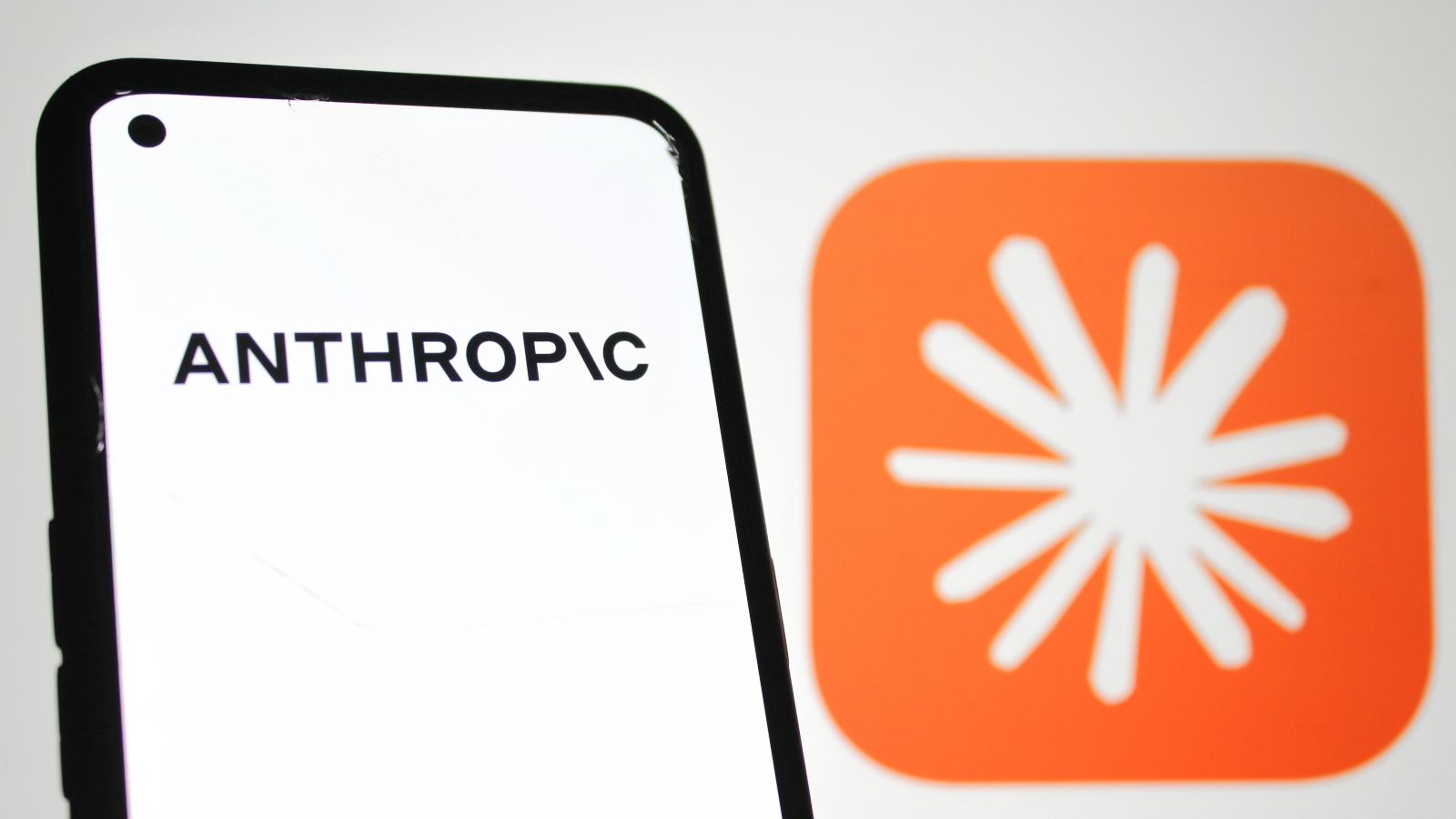 A closeup of a mobile phone displaying the Anthropic logo. Behind in the background is a large Claude logo.