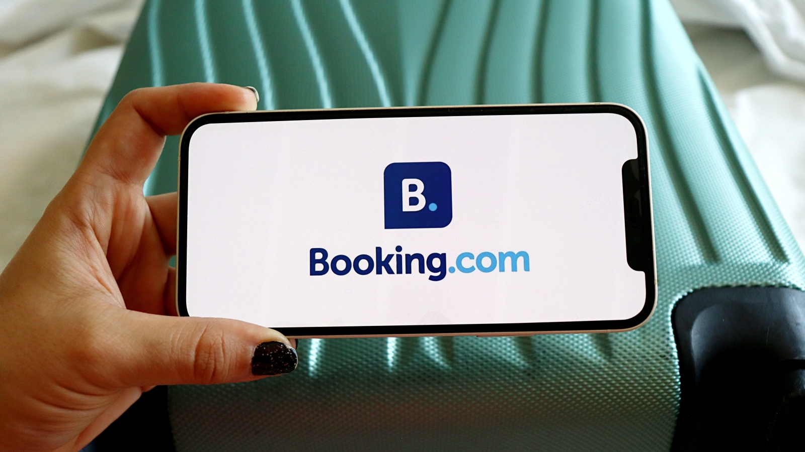 A close up of a smartphone showing the Booking.com logo, being held by someone's hand. In the background is a suitcase on a hotel bed.