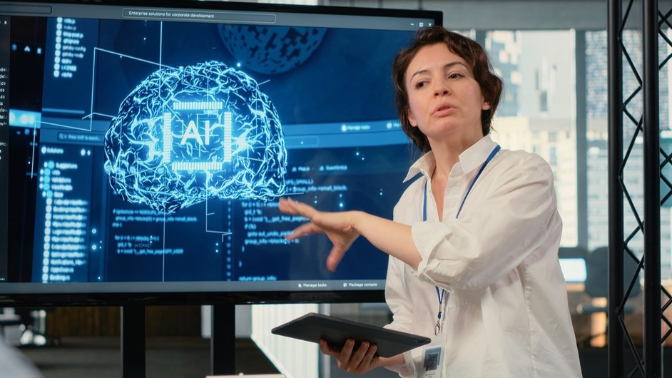 Woman standing and speaking, AI image on the screen next to her.