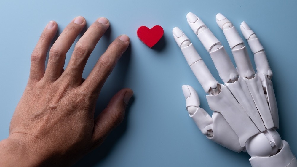 A photo of a human hand next to a robot hand with a small red heart between them.