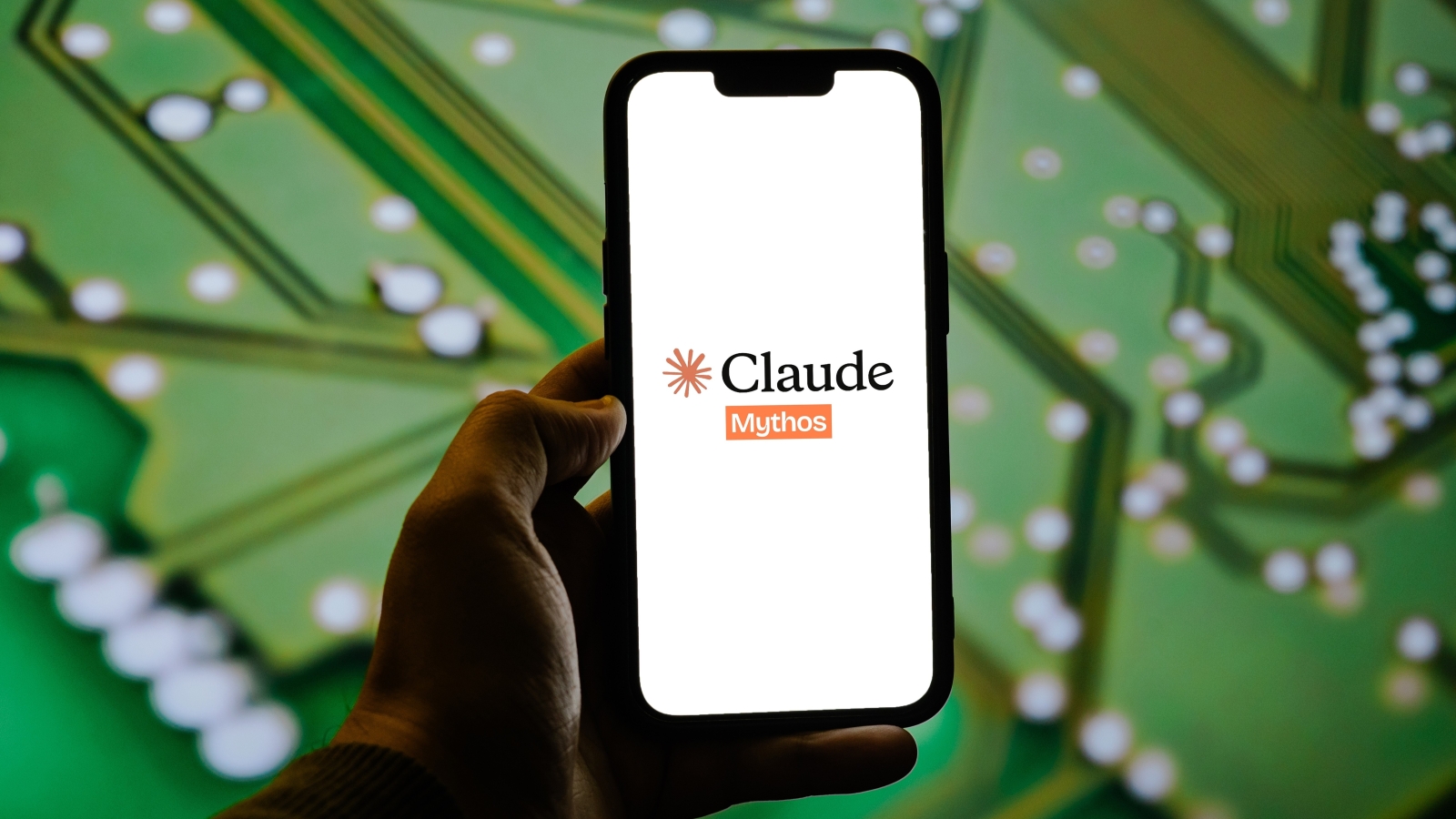 A close up of a smartphone displaying the Claude Mythos logo. In the background, a close up of a computer motherboard.