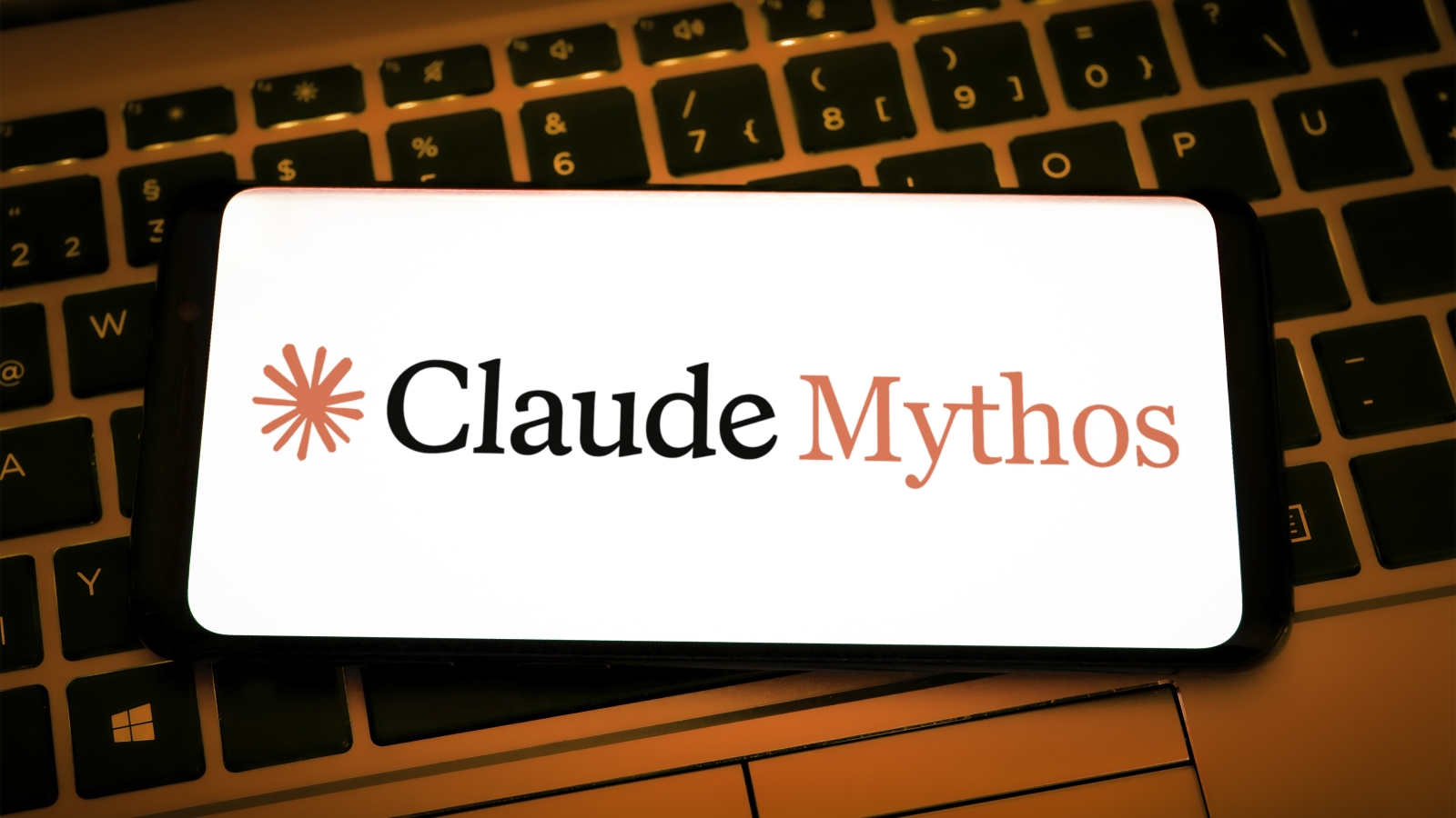 A close up of a smartphone laying on the keyboard of the laptop and displaying a Claude Mythos logo.