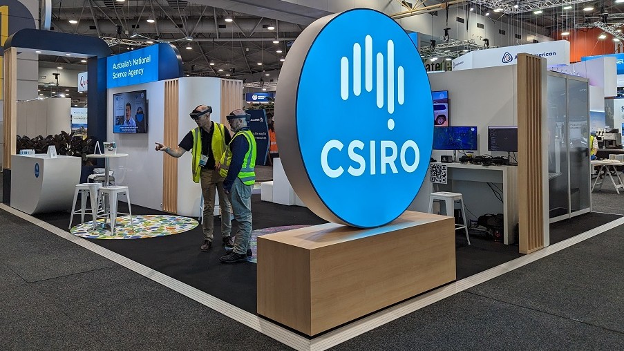 A medium shot of a CSIRO booth at an industry event, with two men in high-vis using virtual reality goggles.