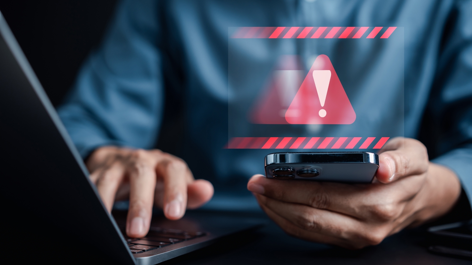 A closeup of a person using a laptop and smartphone, above which hovers a warning sign.