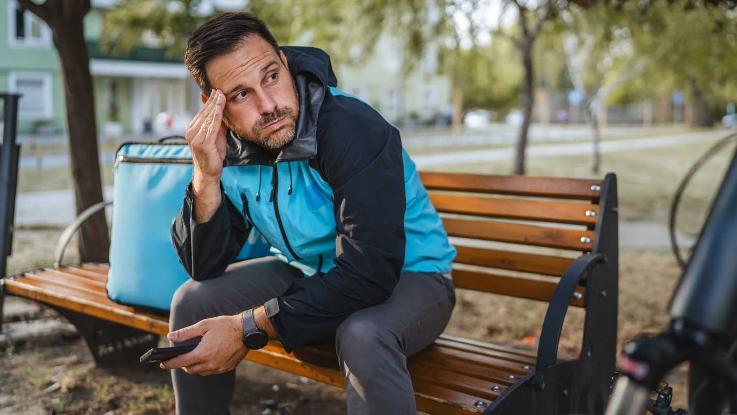 A close up of a male delivery driver sitting on a park bench holding his phone and looking stressed, next to his bike and delivery capsule.