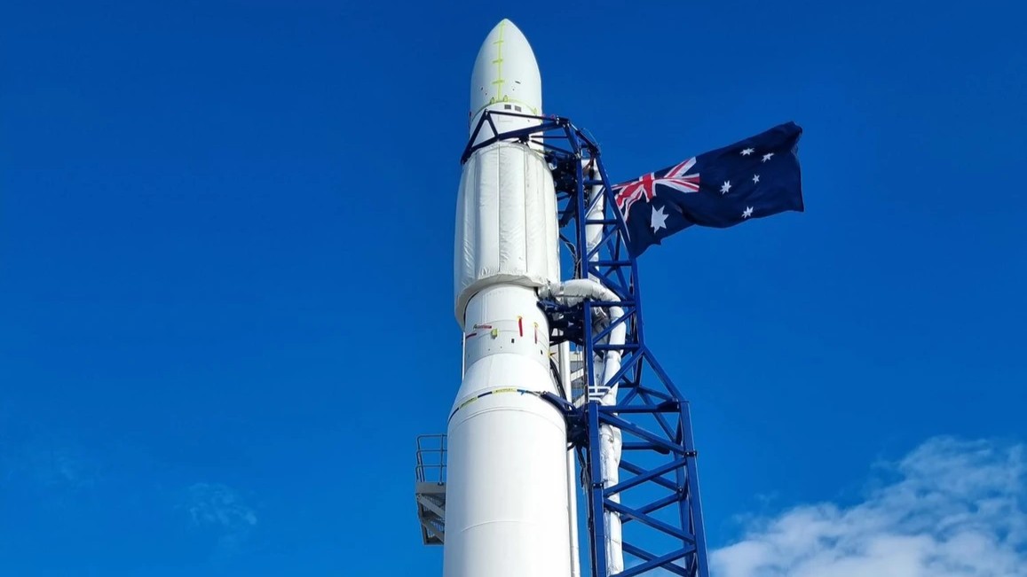 A close up of the top of an orbital rocket, with an Australian flag flying next to it and clear sky in the background.