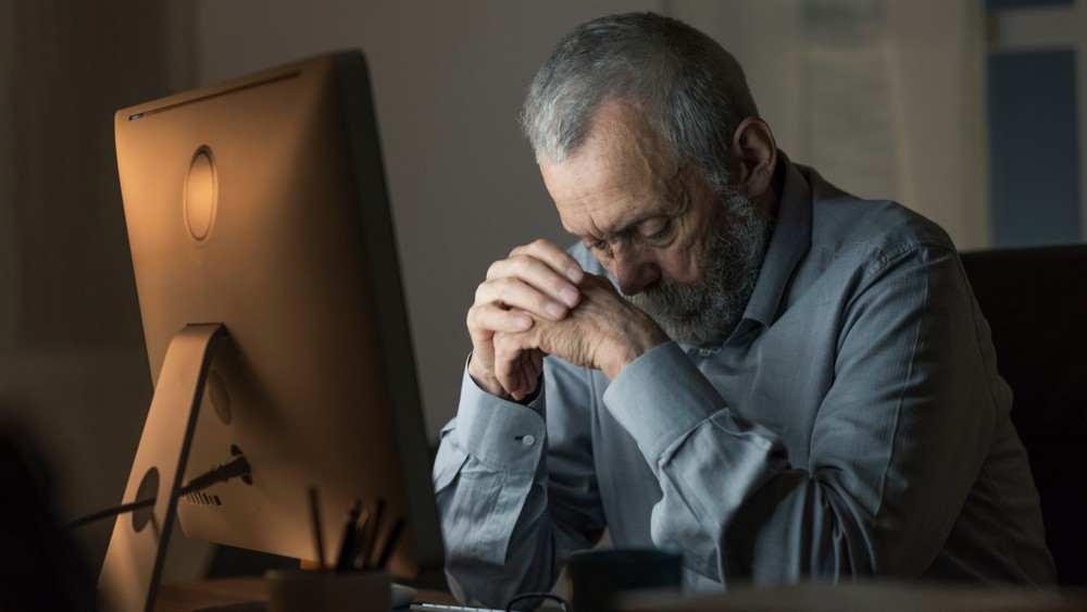 Older man sitting in front of a computer in the dark, looking downwards.