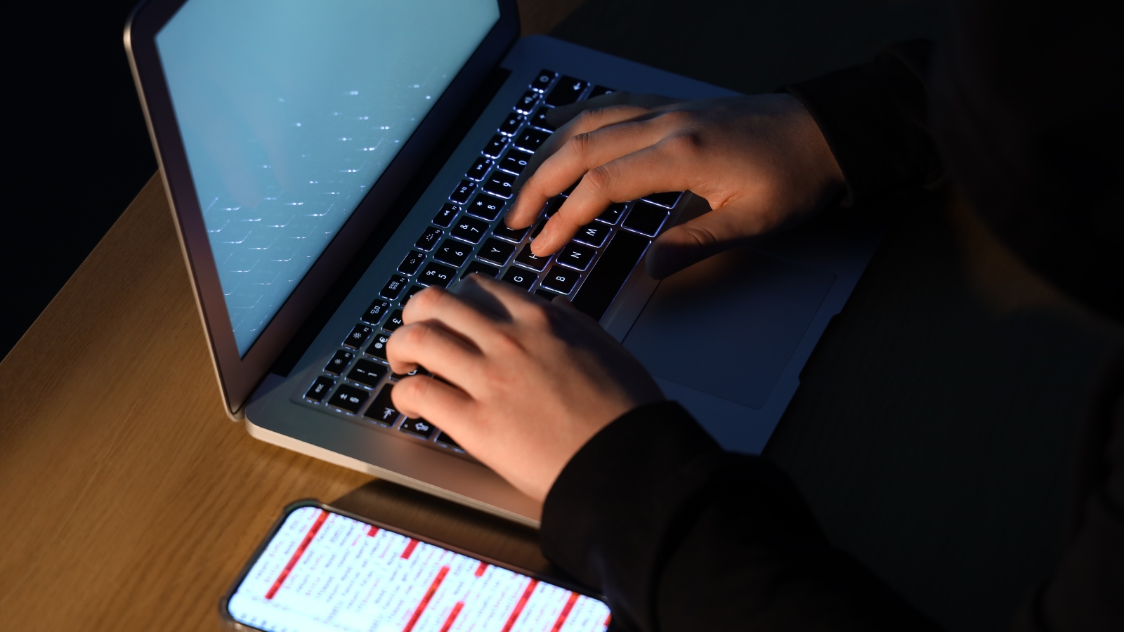 A close up of a man's hands as he uses a laptop and smartphone in a dark room.