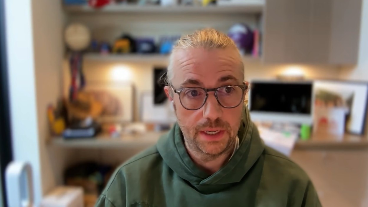 A closeup of Mike Cannon-Brookes sitting in his office, wearing a hoodie and glasses, and speaking to camera